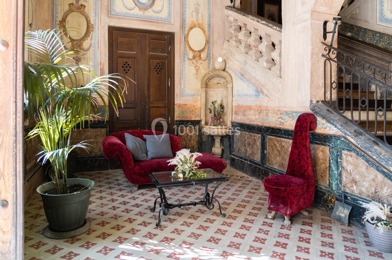 Salon avec fauteuils rouges, table basse en verre, plante en pot et fontaine décorative dans un intérieur ancien.