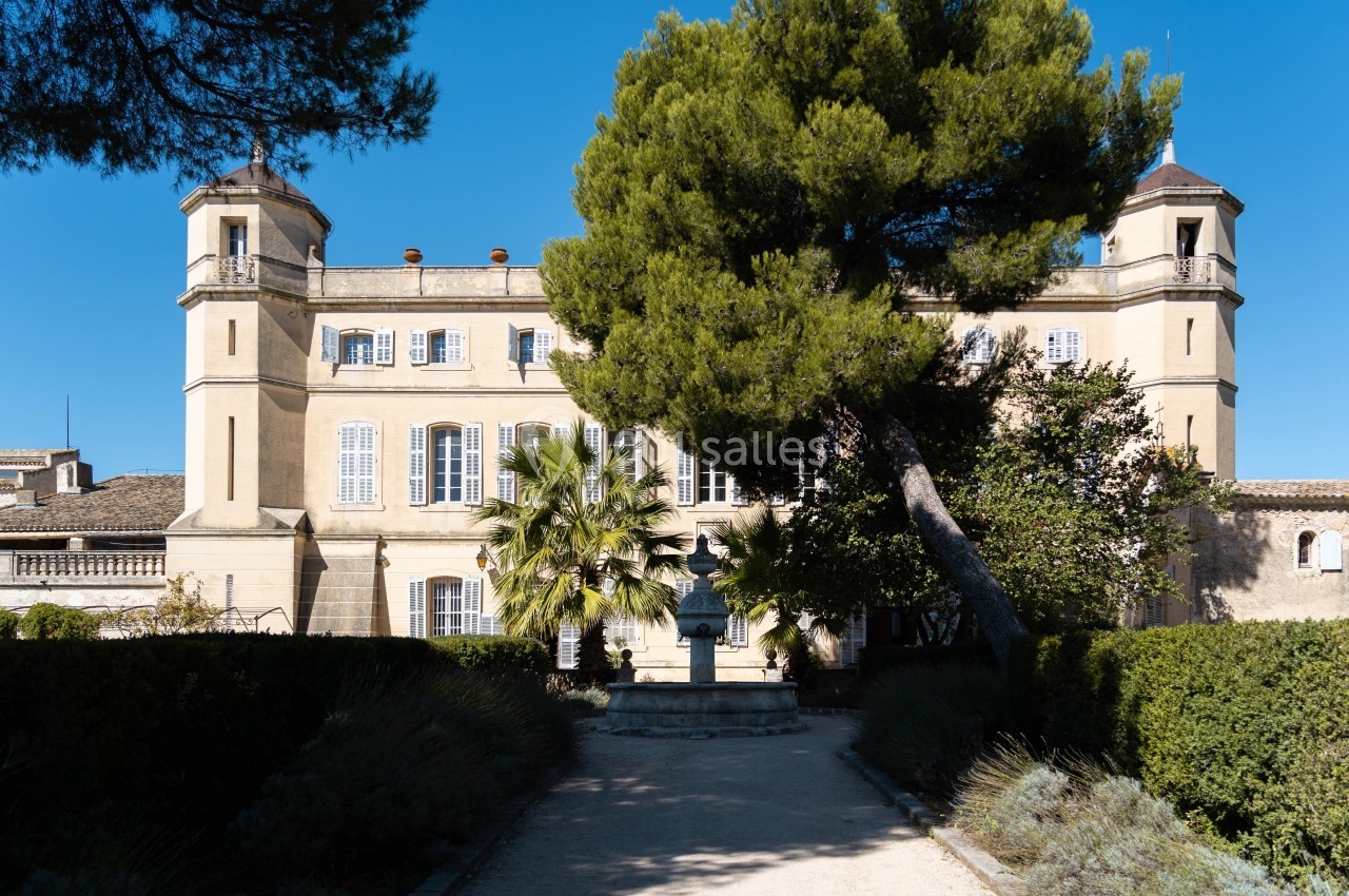 Façade d'un château beige entouré d'arbres et de végétation sous un ciel bleu clair.