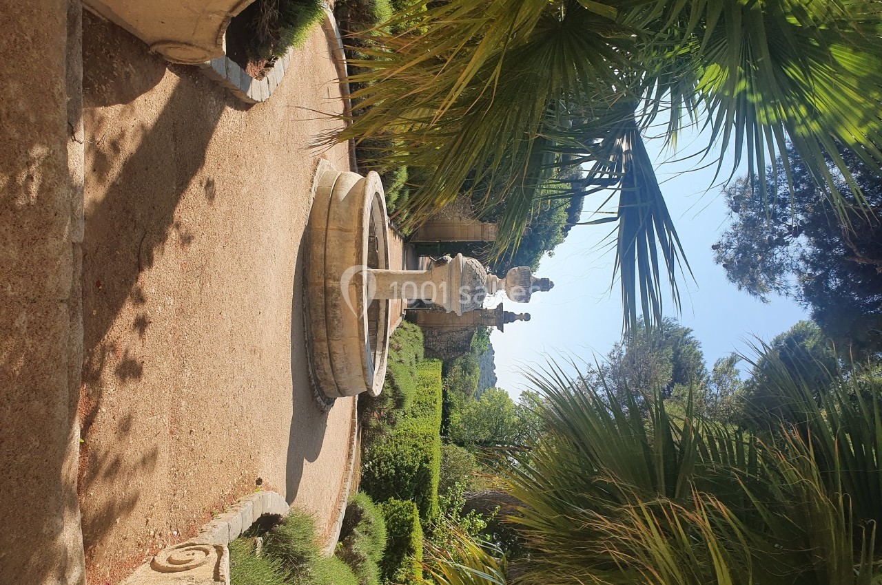 Fontaine en pierre entourée de végétation méditerranéenne dans un jardin ensoleillé avec des palmiers et des allées…