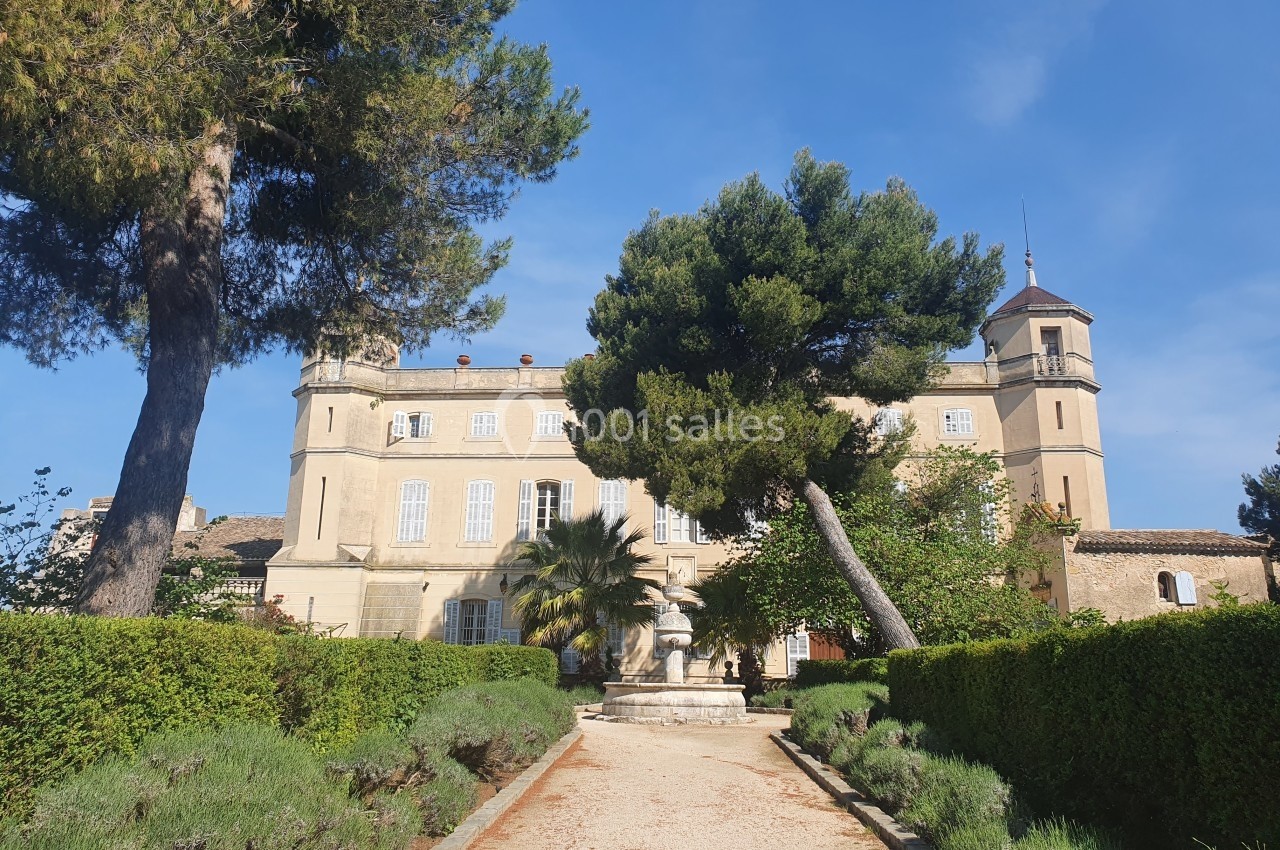 Façade d'un château entouré de jardins, avec allée bordée de buissons et statue au centre, sous un ciel bleu.