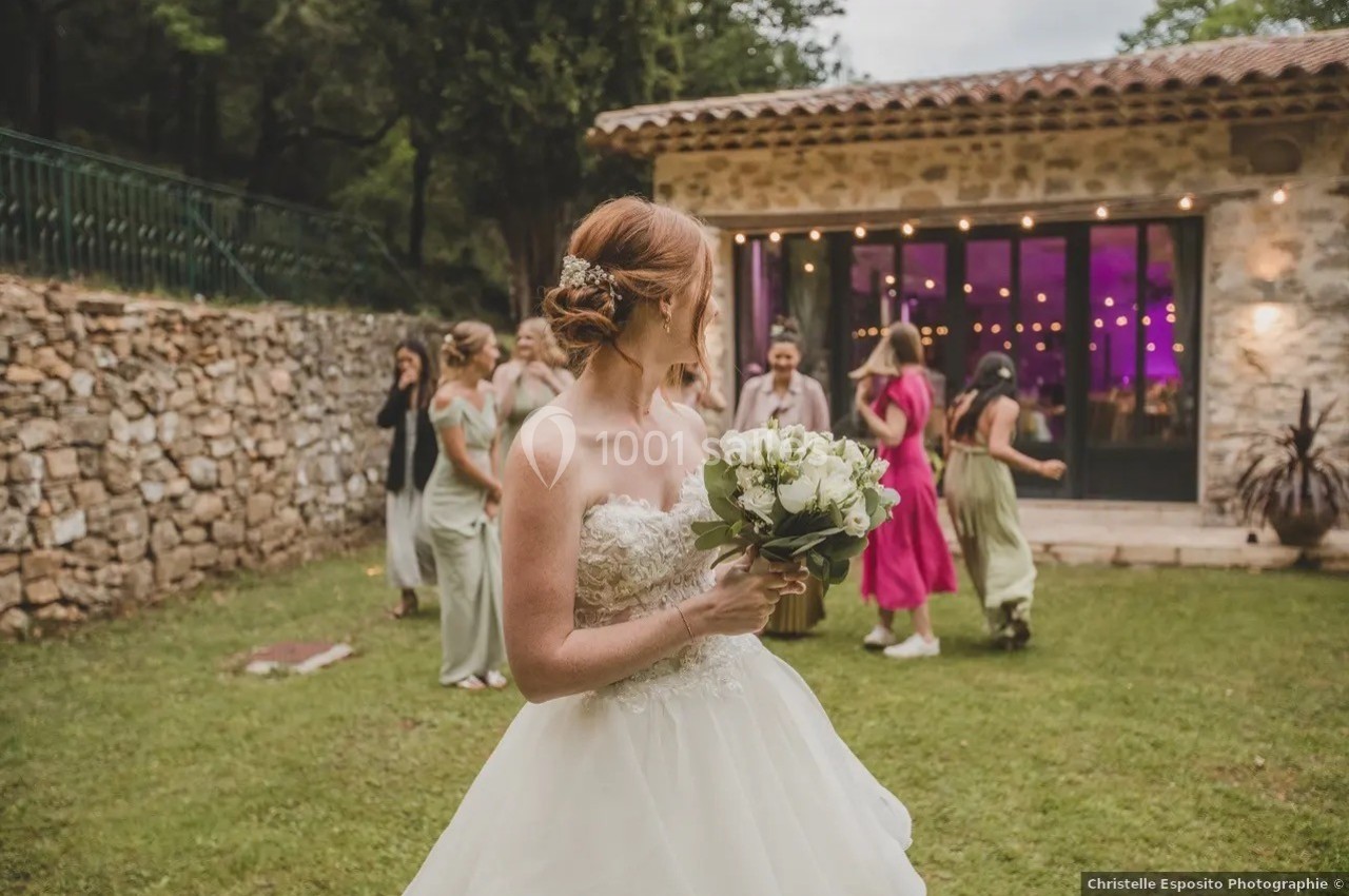 Une mariée en robe blanche tient un bouquet, entourée d'invités dans un jardin devant une salle éclairée.