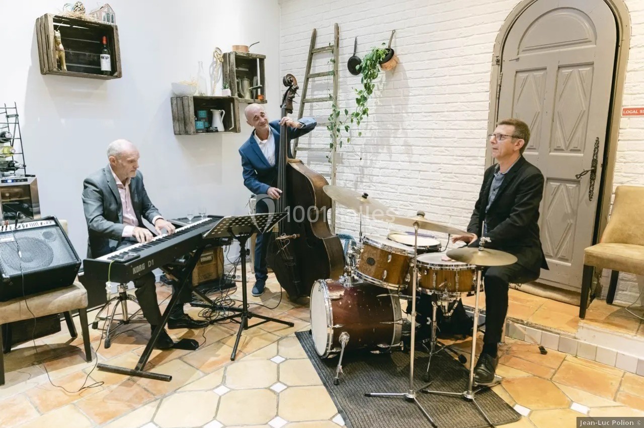 Trois musiciens jouent du piano, de la contrebasse et de la batterie dans une pièce au décor chaleureux.