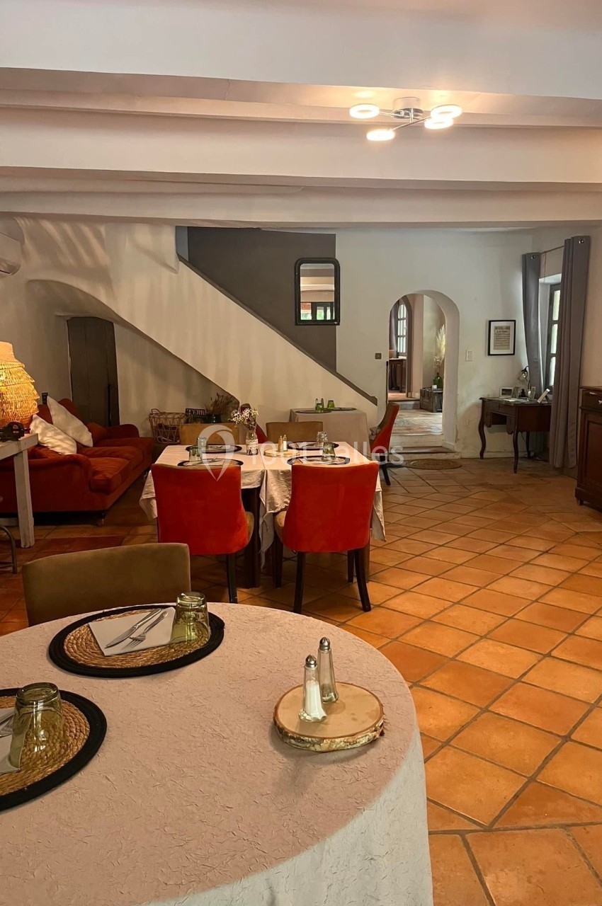 Salle à manger chaleureuse avec tables dressées, sol en terre cuite, canapé rouge et escalier menant à l'étage.