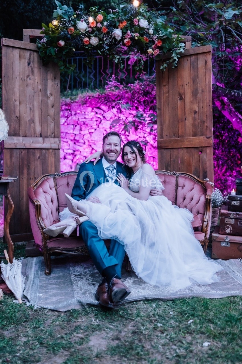 Un couple assis sur un canapé vintage devant une arche fleurie, dans un décor extérieur éclairé en rose.
