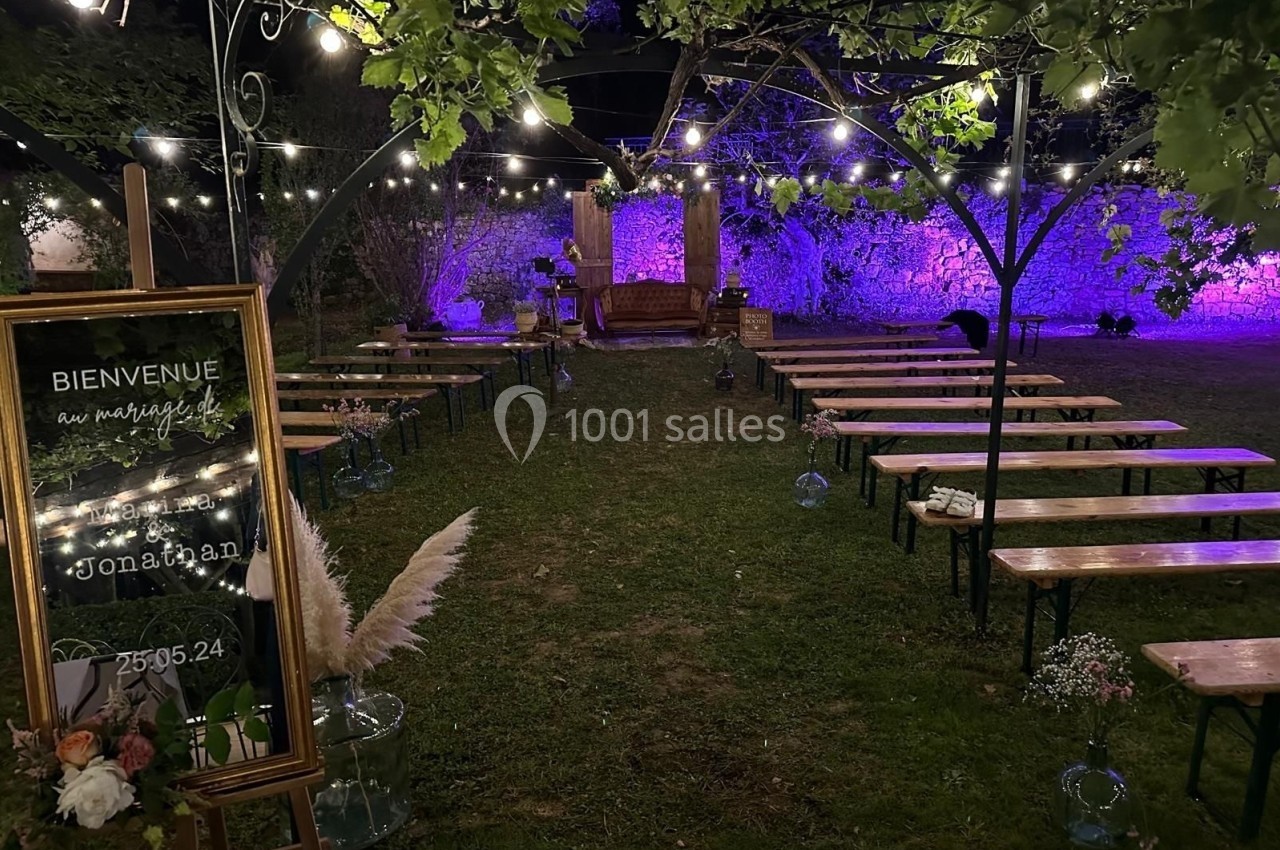 Allée décorée pour une cérémonie en extérieur, avec bancs en bois, guirlandes lumineuses et panneau de bienvenue.