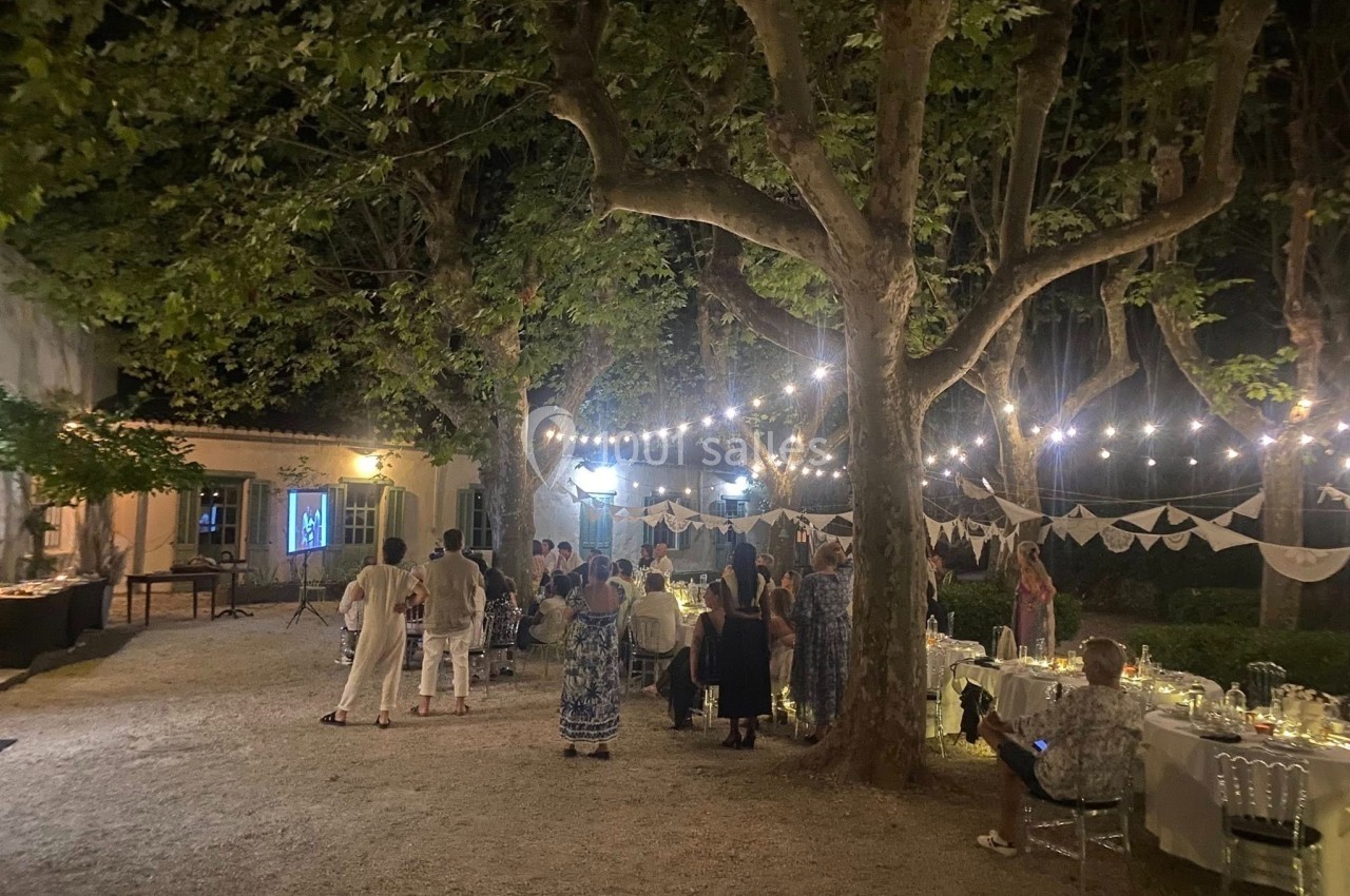 Groupe de personnes rassemblées en soirée dans un jardin éclairé par des guirlandes lumineuses, tables dressées à proximité.