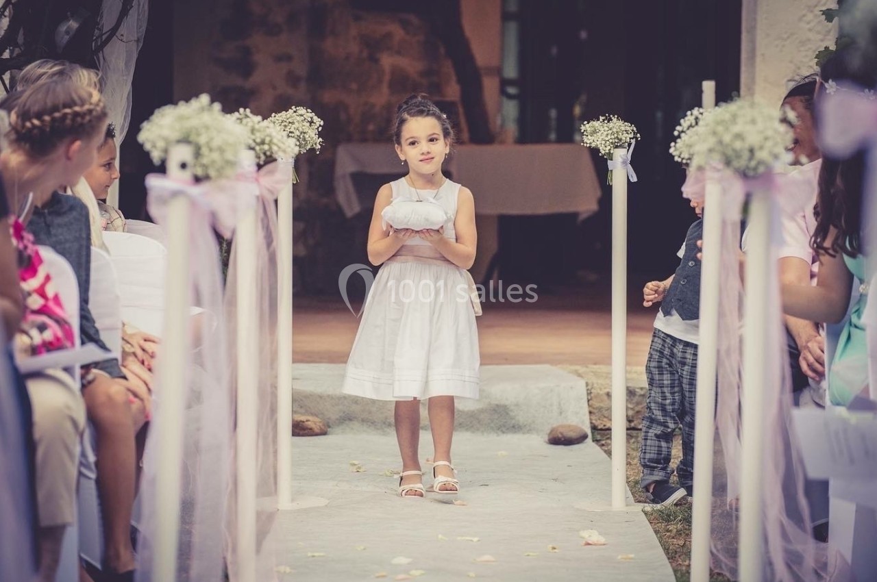 Une jeune fille en robe blanche porte un coussin sur une allée décorée lors d'une cérémonie en plein air.