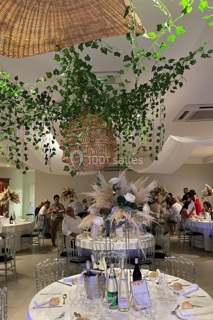 Salle de réception décorée avec des plantes suspendues, des tables dressées et des invités assis.