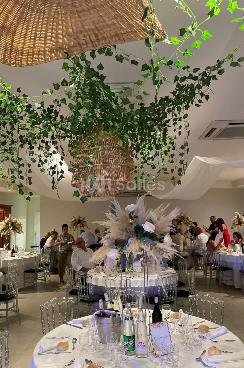 Salle de réception décorée avec des plantes suspendues, des tables dressées et des invités assis.