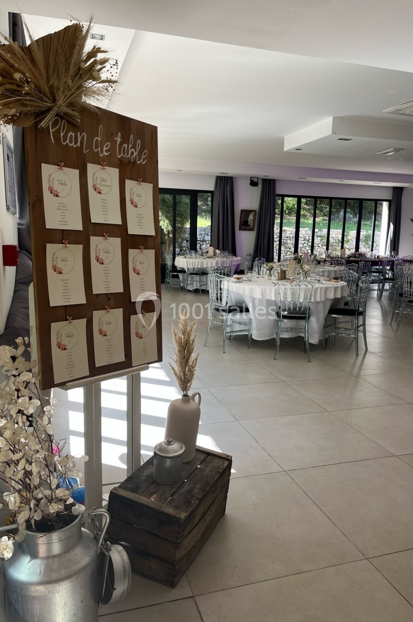 Salle de réception décorée avec des tables dressées et un plan de table affiché sur un panneau en bois.