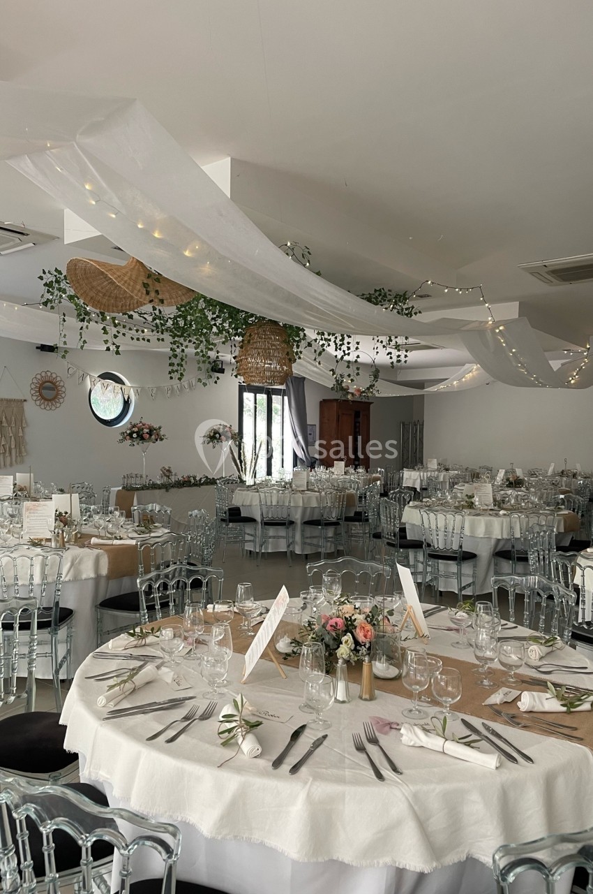 Salle de réception décorée pour un mariage, avec tables rondes dressées, nappes blanches et guirlandes lumineuses suspendues.