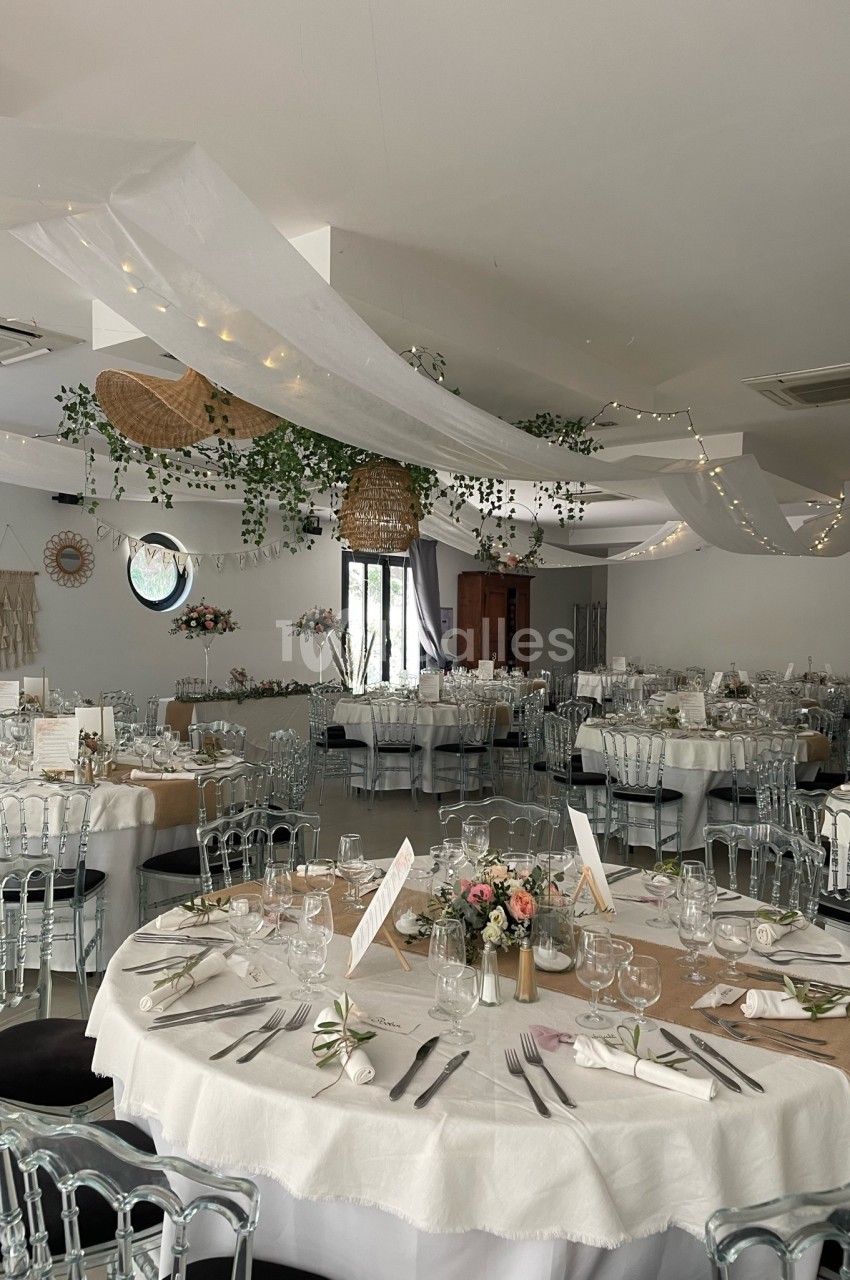 Salle de réception décorée pour un mariage, avec tables rondes dressées, nappes blanches et guirlandes lumineuses suspendues.