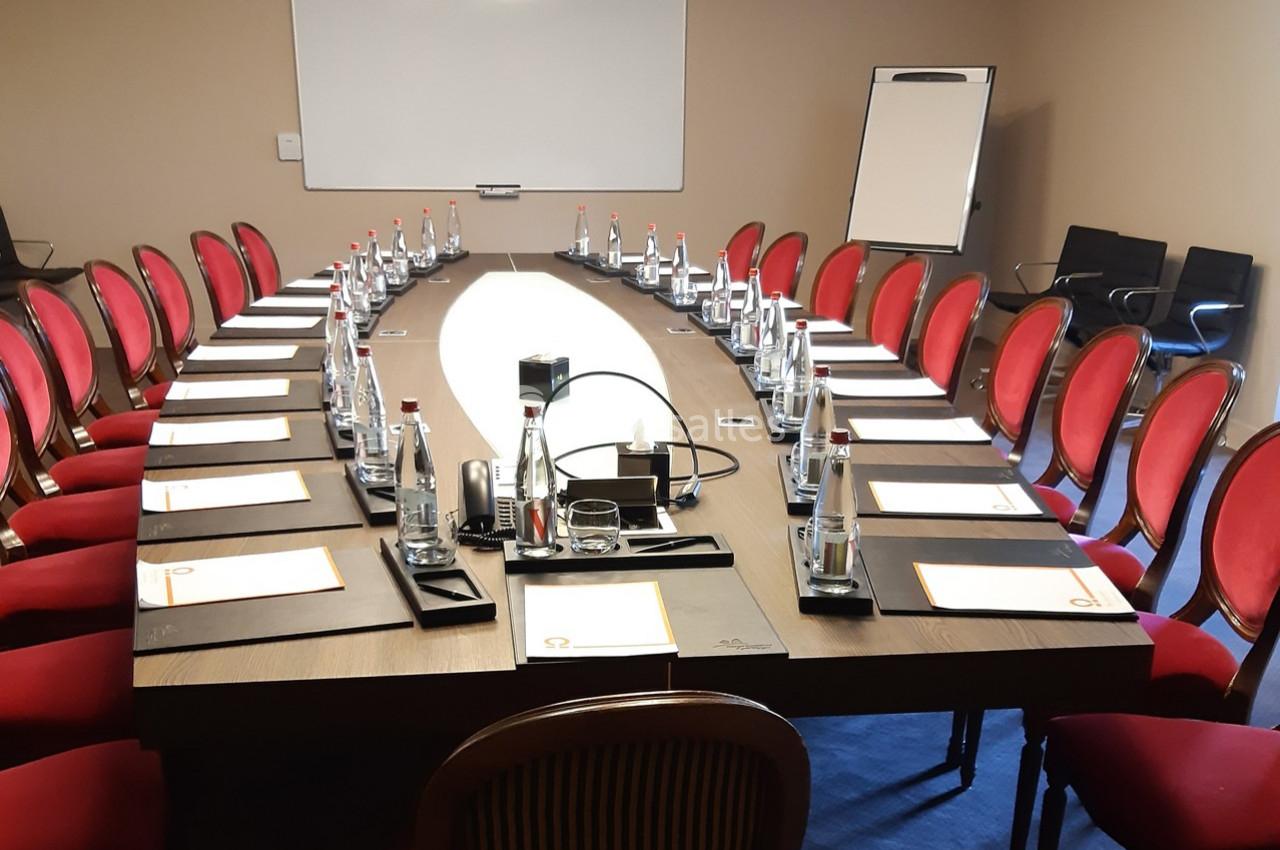 Salle de réunion avec une grande table ovale, chaises rouges, documents, bouteilles d'eau et tableau blanc en arrière-plan.