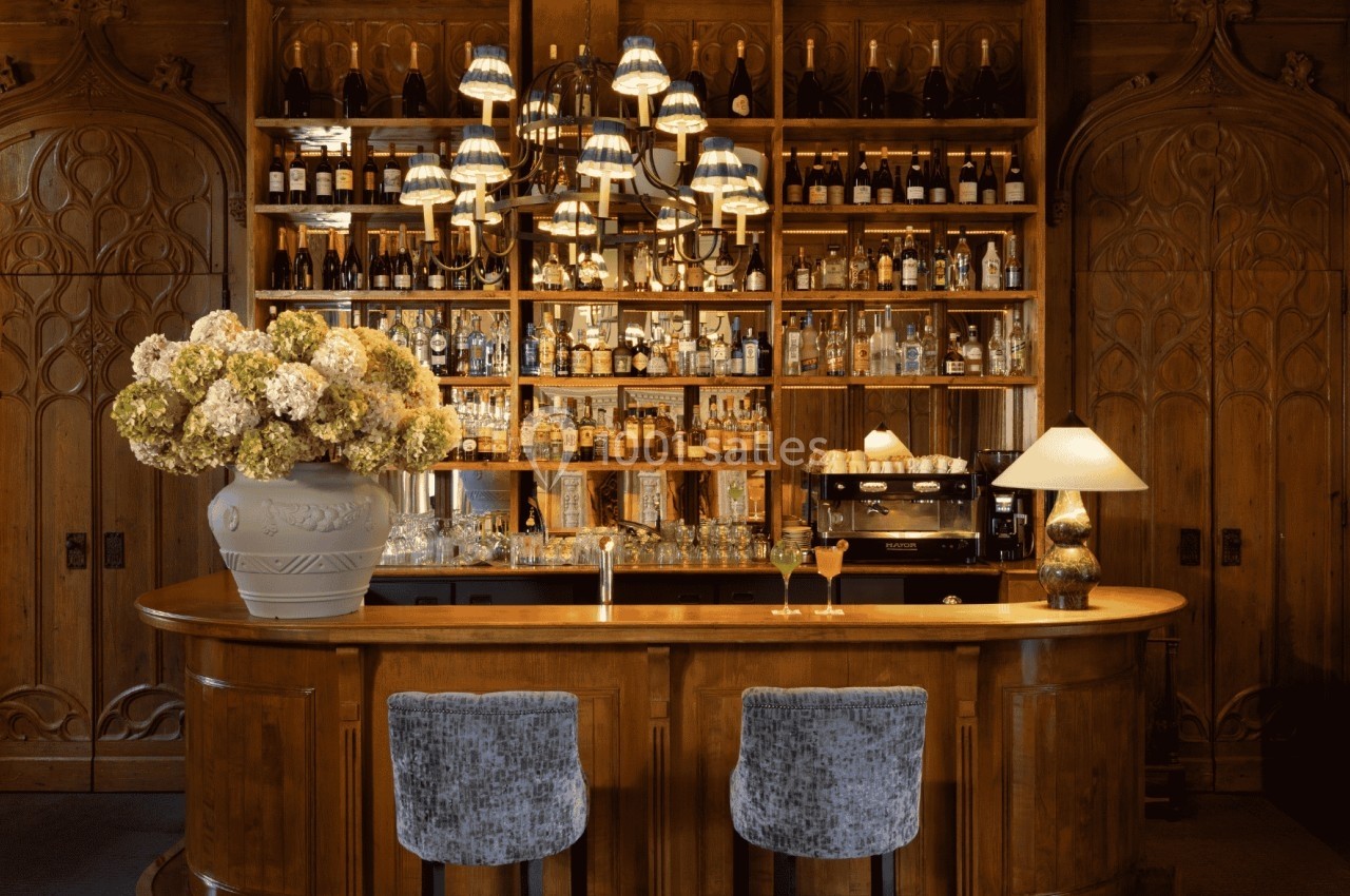 Bar en bois élégant avec étagères remplies de bouteilles, deux tabourets, un vase de fleurs et des luminaires suspendus.