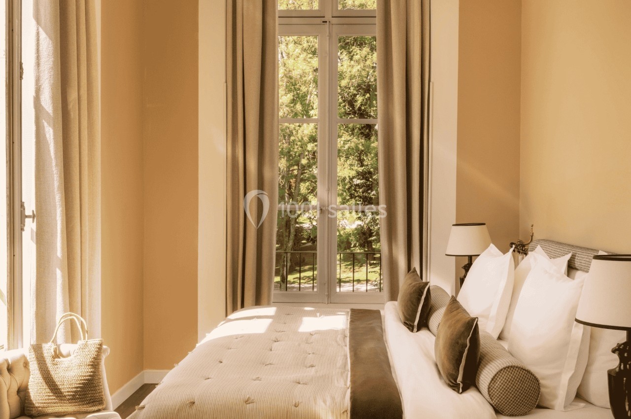 Chambre lumineuse avec un grand lit, des coussins, des rideaux beiges et une fenêtre donnant sur un jardin verdoyant.