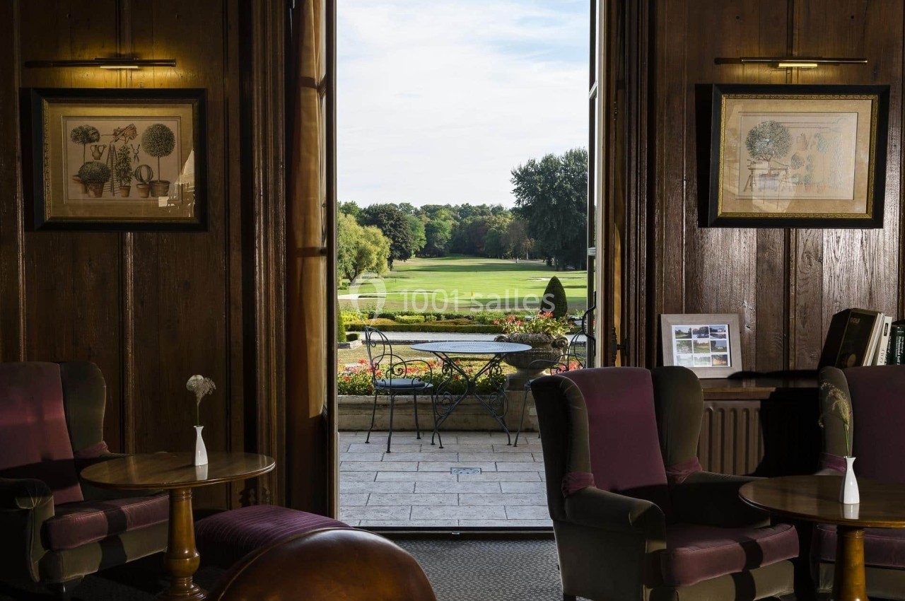 Vue sur un jardin verdoyant depuis un salon élégant avec fauteuils, boiseries et tableaux encadrés.