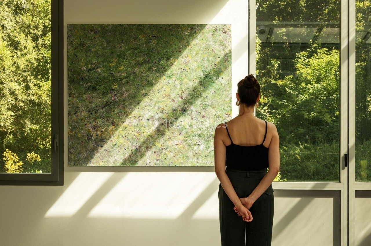 Une femme observe un tableau abstrait dans une pièce lumineuse avec de grandes fenêtres donnant sur un jardin verdoyant.