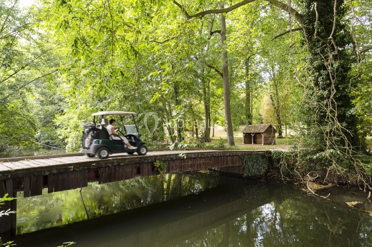 Un golf cart traverse un pont en bois au-dessus d'une rivière entourée d'arbres et de verdure.