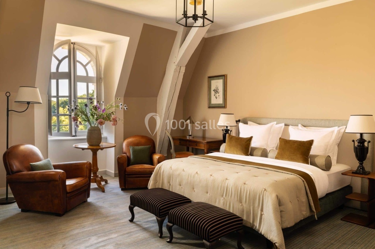 Chambre lumineuse avec lit double, fauteuils en cuir, table basse, fenêtres cintrées et décoration chaleureuse.
