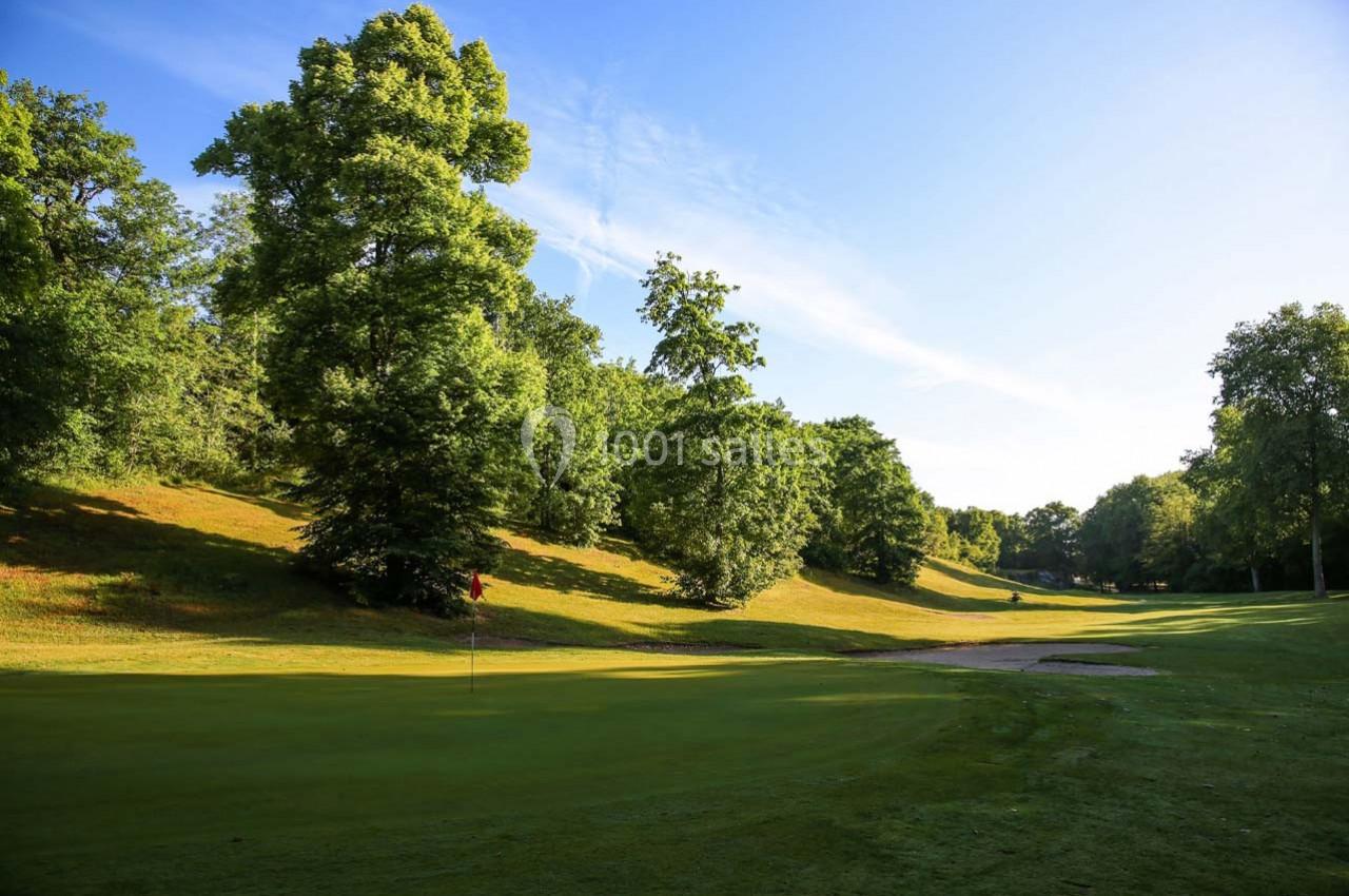 Paysage d'un terrain de golf avec un drapeau rouge, entouré d'arbres et baigné de lumière naturelle.