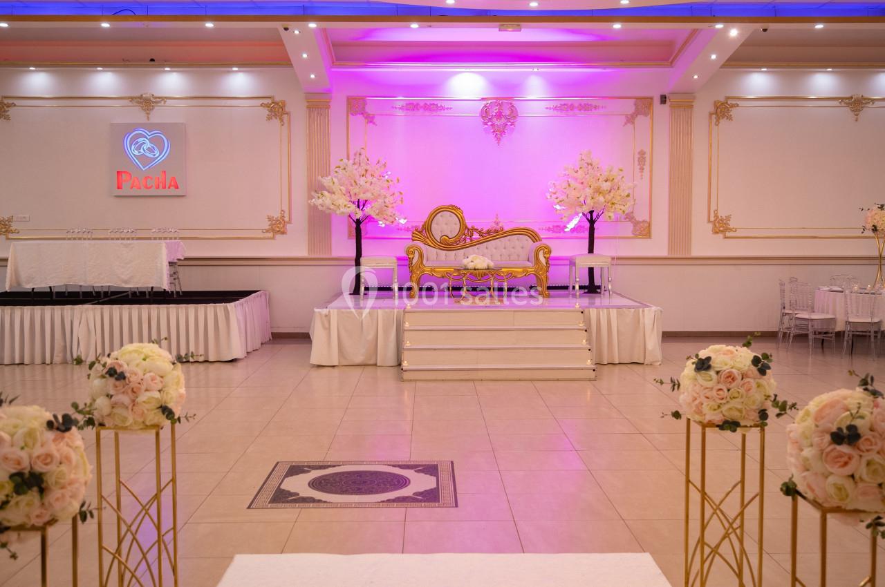 Salle de réception décorée avec un canapé doré sur une estrade, éclairage rose et arrangements floraux.