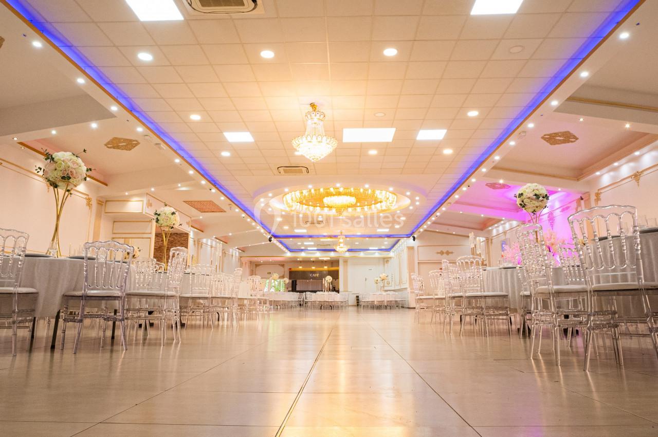 Salle de réception élégante avec tables dressées, chaises transparentes et lustres illuminant le plafond décoré.