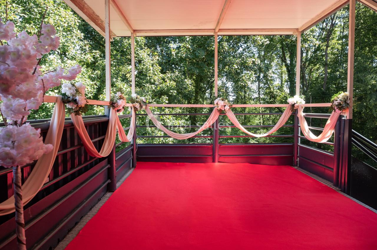 Terrasse décorée avec des fleurs et des rubans, sol recouvert d’un tapis rouge, entourée d’arbres verdoyants.