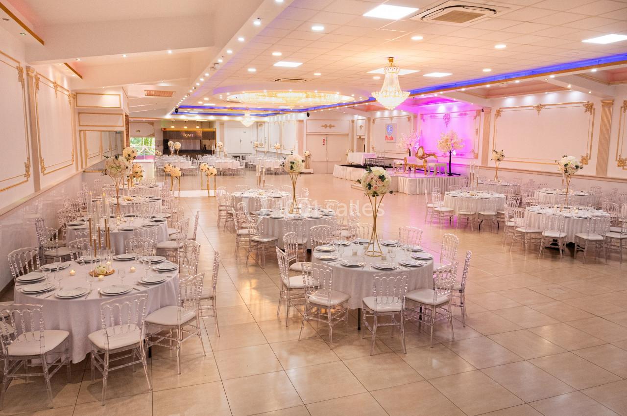 Salle de réception élégante avec tables rondes dressées, chaises transparentes et éclairage décoratif.