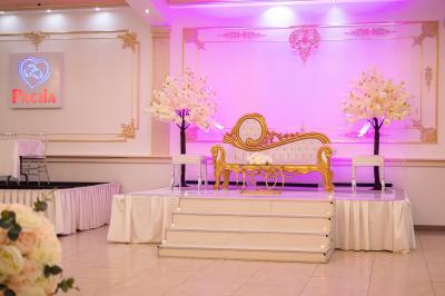 Salle de réception décorée avec un canapé doré, des arbres fleuris et un éclairage rose sur une estrade blanche.