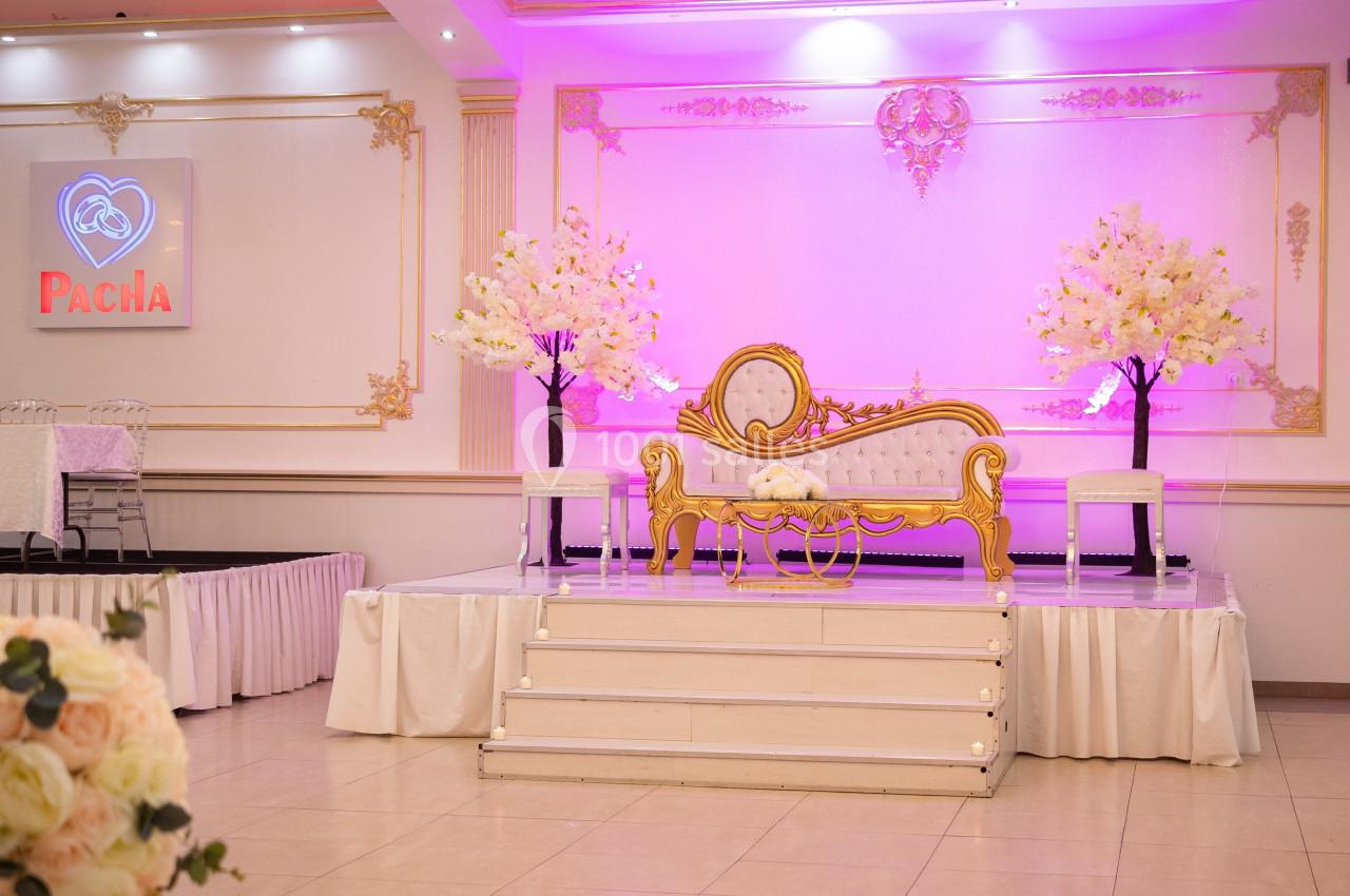 Salle de réception décorée avec un canapé doré, des arbres fleuris et un éclairage rose sur une estrade blanche.