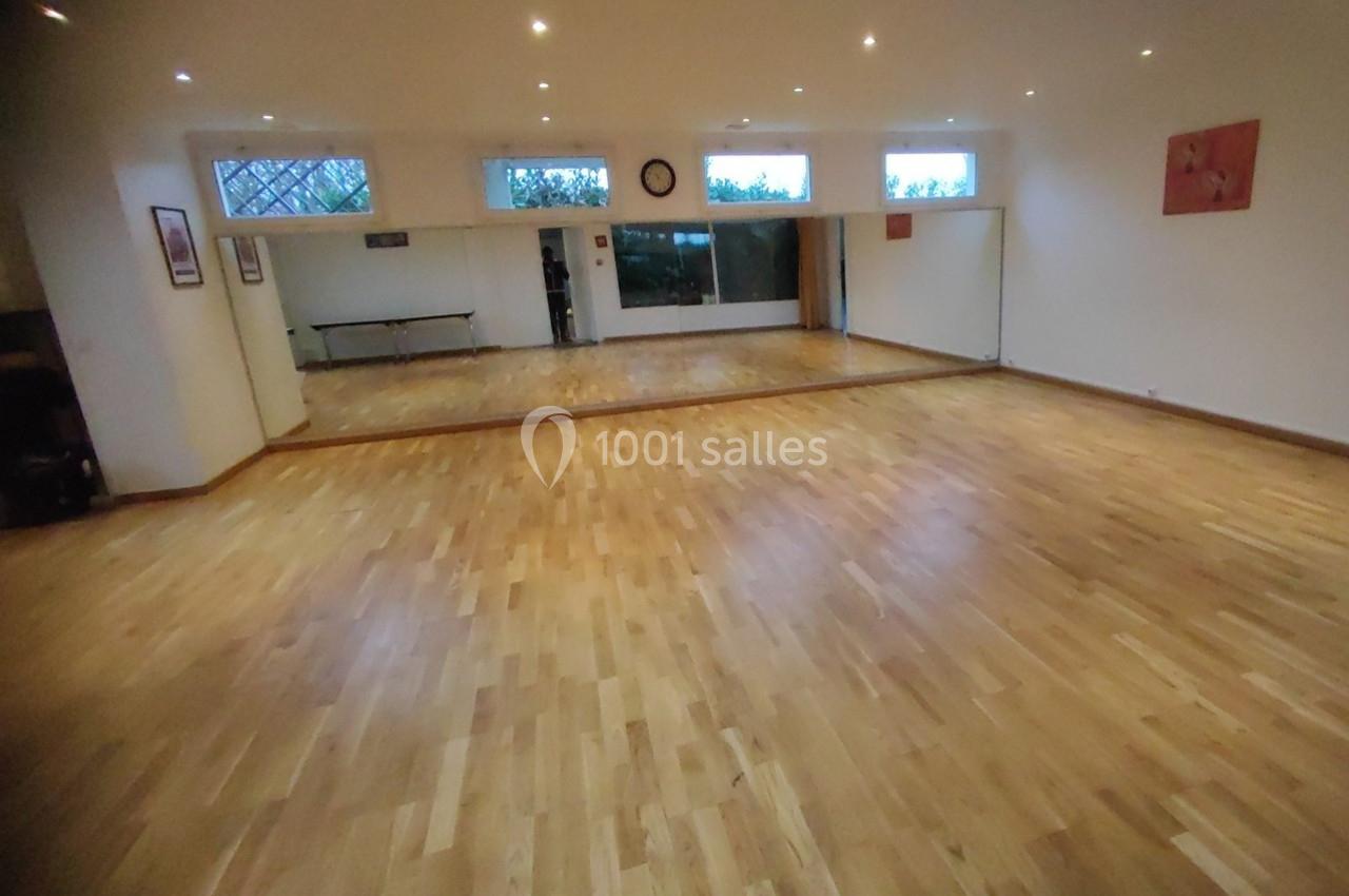 Salle lumineuse avec parquet en bois clair, murs blancs, miroirs sur un mur et fenêtres en hauteur.