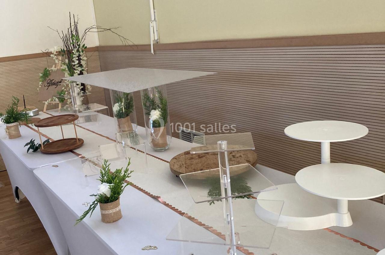 Table décorée pour un buffet, avec supports vides, plantes en pot, branches et nappes blanches dans une salle lumineuse.