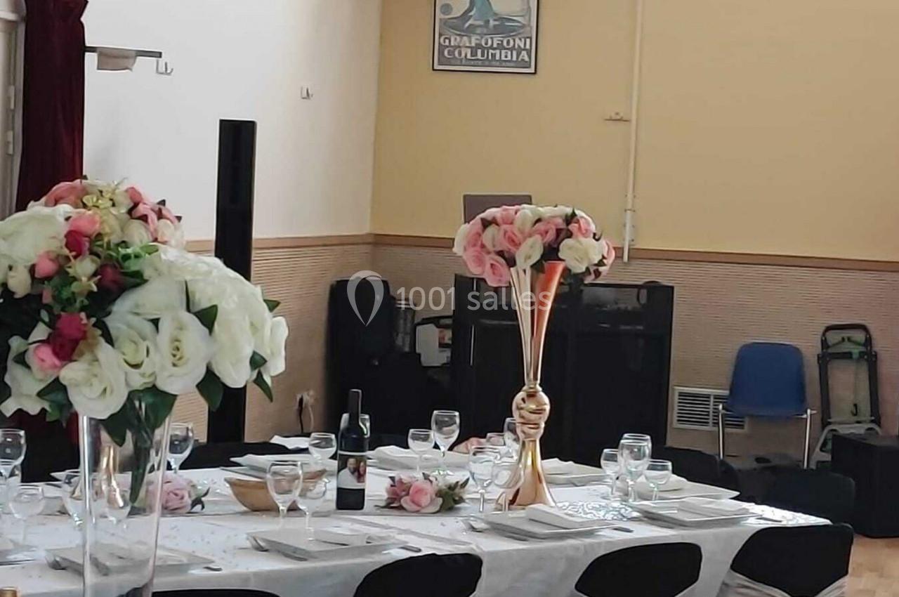 Table dressée pour un événement avec nappes blanches, verres, assiettes et bouquets de fleurs en centre de table.