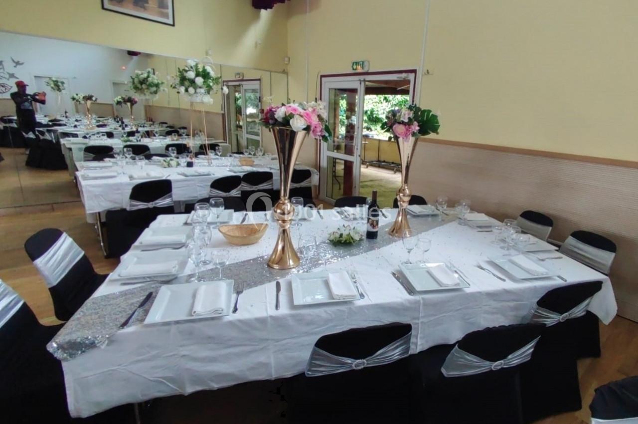 Salle décorée pour un événement avec tables dressées, nappes blanches, centres de table fleuris et chaises noires ornées.