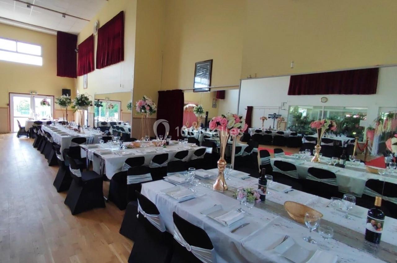 Salle de réception décorée avec des tables alignées, nappes blanches, chaises noires et arrangements floraux roses.