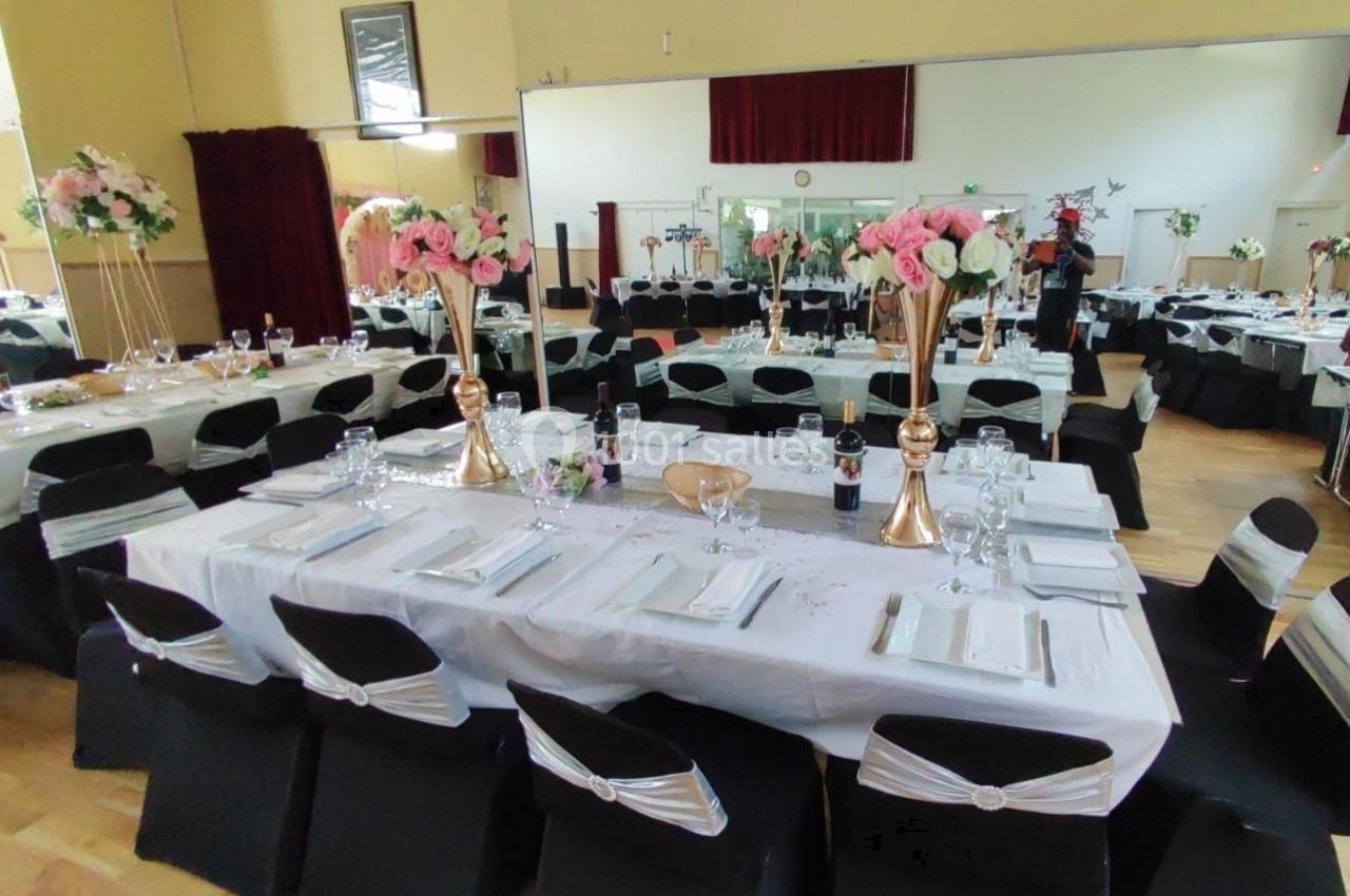 Salle de réception décorée avec des tables dressées, nappes blanches, chaises noires et arrangements floraux.