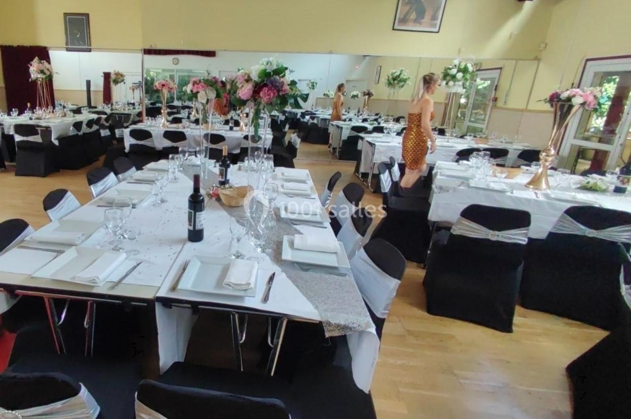 Salle décorée pour un événement avec tables dressées, nappes blanches, chaises noires et arrangements floraux.