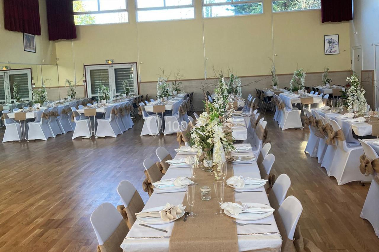 Salle de réception décorée avec des tables alignées, nappes blanches, chemins de table en toile de jute et bouquets floraux.