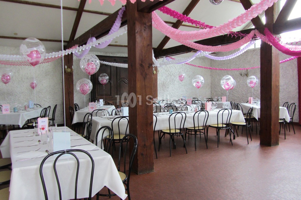 Salle décorée pour une fête avec des tables dressées, des guirlandes roses et blanches, et des ballons suspendus.