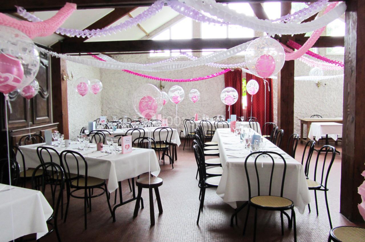 Salle décorée pour une réception avec des tables dressées, des chaises noires et des guirlandes roses suspendues.