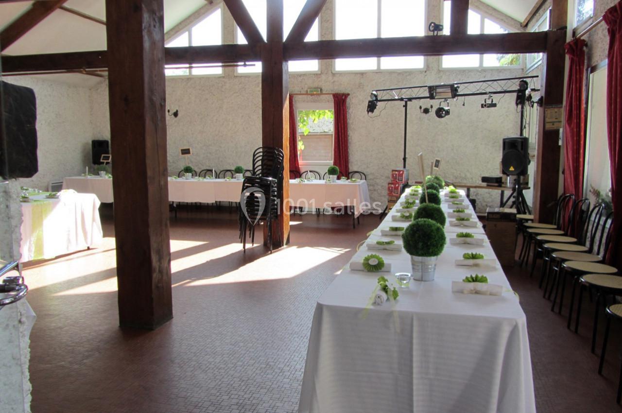 Salle lumineuse avec tables dressées, chaises empilées, poutres en bois et équipement audio en arrière-plan.