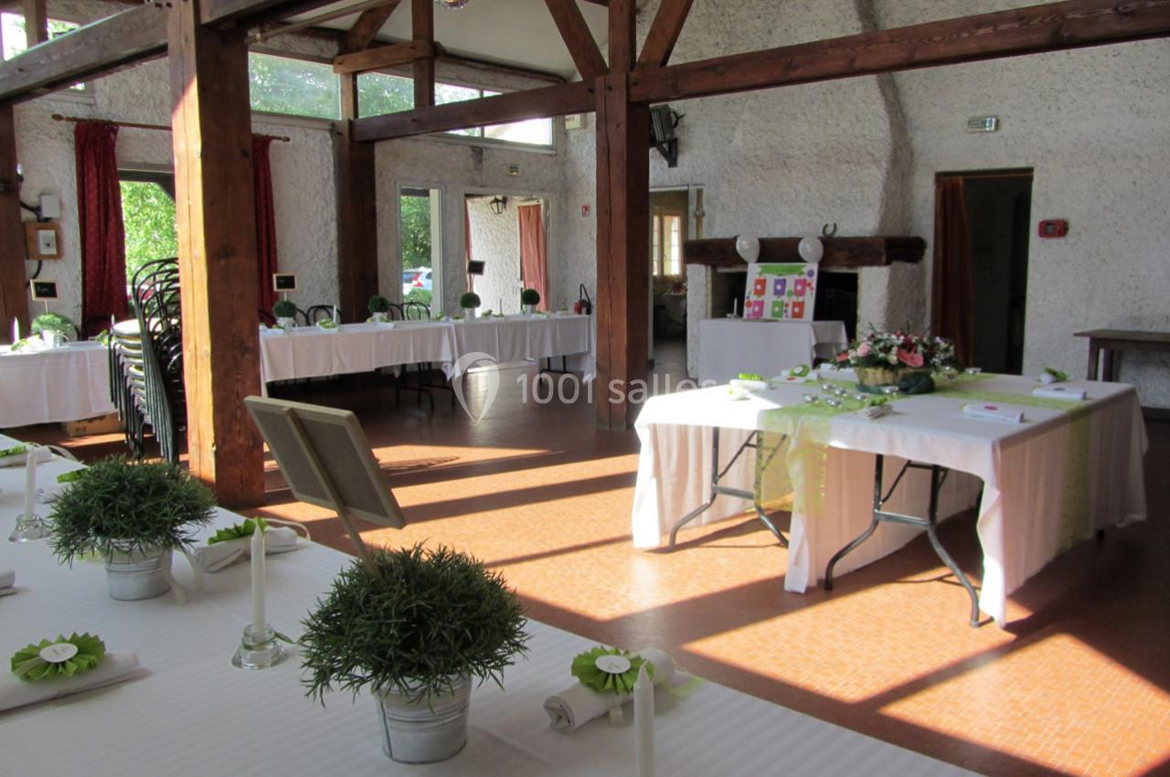 Salle lumineuse décorée pour un événement, avec tables dressées, nappes blanches et arrangements floraux.