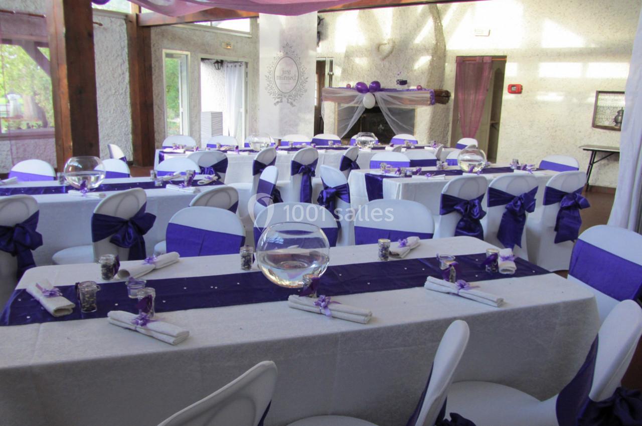Salle décorée pour un événement avec tables dressées, nappes blanches, chemins de table violets et chaises assorties.
