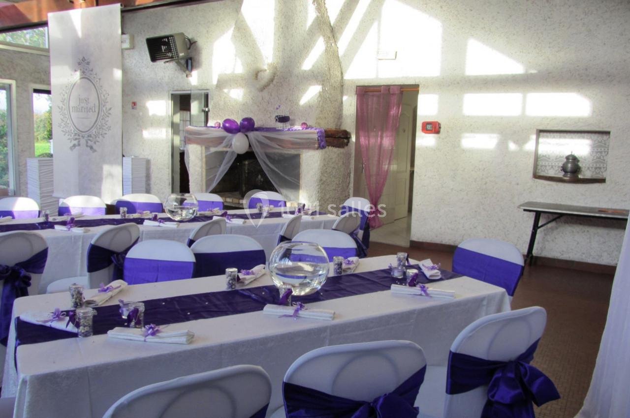 Salle décorée pour un événement avec tables dressées, nappes blanches, chemins de table violets et chaises assorties.