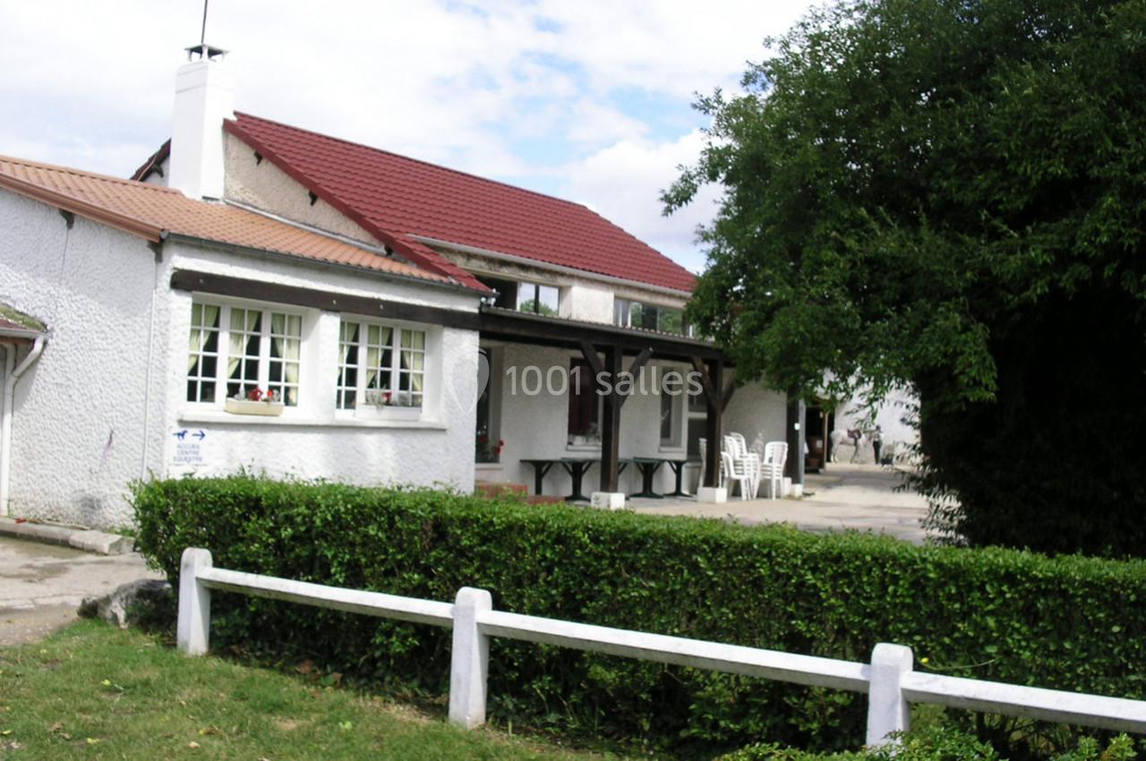 Maison avec façade blanche, toit rouge et terrasse équipée de tables et chaises, entourée de verdure et d'une haie.