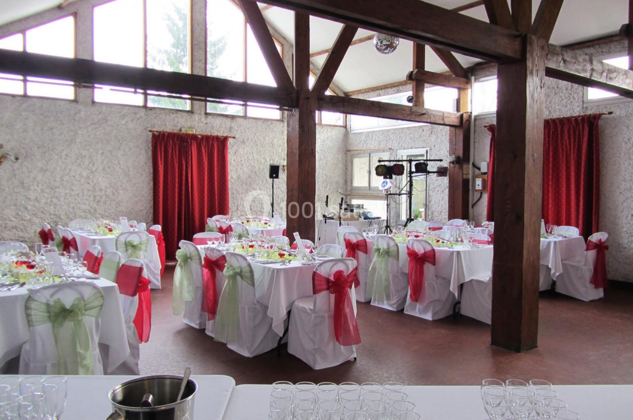 Salle décorée pour un événement avec des tables dressées, nappes blanches et chaises ornées de nœuds rouges et verts.