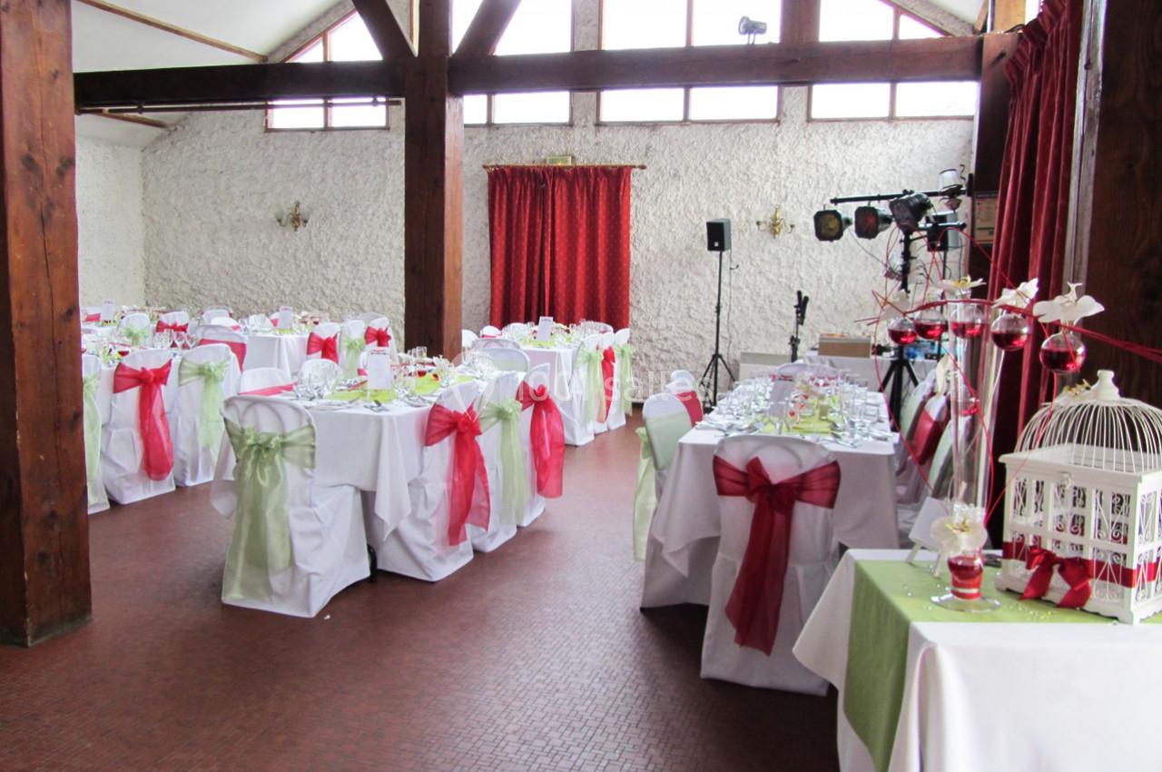 Salle de réception décorée avec des tables dressées, nappes blanches et chaises ornées de nœuds rouges et verts.