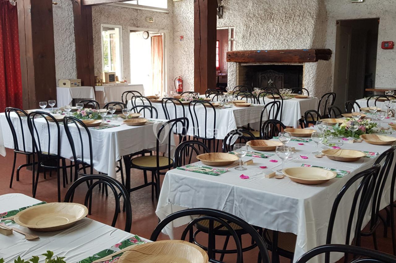 Salle de réception avec tables dressées, nappes blanches, assiettes en bois et décorations florales.