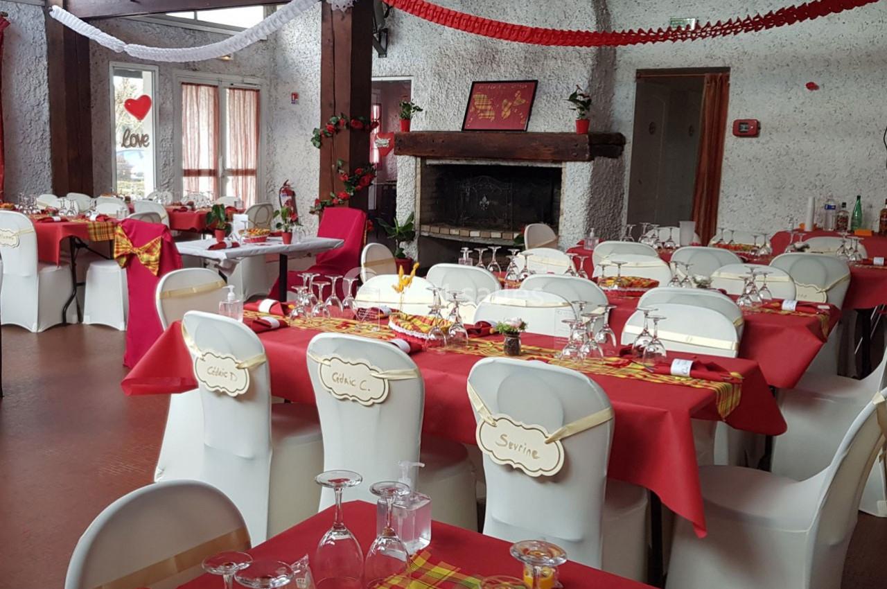 Salle décorée pour un événement, avec des tables dressées, nappes rouges et jaunes, et chaises blanches ornées.