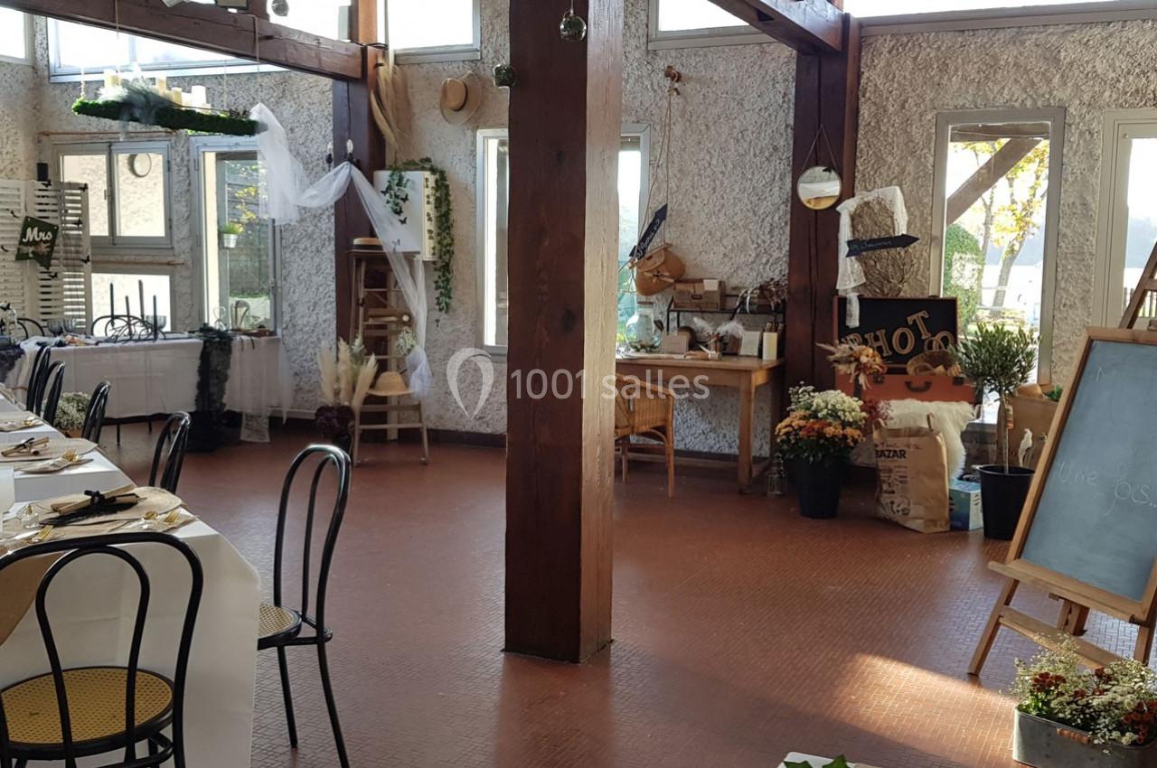 Salle lumineuse décorée pour un événement, avec tables dressées, plantes et éléments rustiques en arrière-plan.